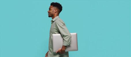 A solo studio portrait featuring an AfricanAmerican man in casual attire holding a closed laptop while walking alone against a soft light blue backdrop photo