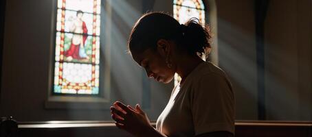 An African American woman kneels in prayer with sunlight streaming through stainedglass windows during a profoundly spiritual moment at church photo