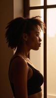 African American young woman with an Afro stands at a window bathed in golden light reflectively gazes out photo