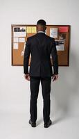An AfricanAmerican man is depicted from behind as he stands before a cluttered corkboard filled with notes photo