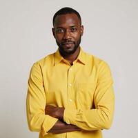 An African American man poses in a bright studio portrait wearing a yellow shirt and crossing his hands against a white backdrop photo