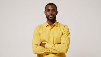 An African American man poses in a bright studio portrait wearing a yellow shirt and crossing his hands against a white backdrop photo