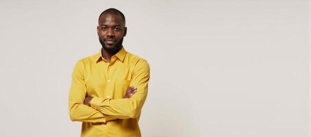 An African American man poses in a bright studio portrait wearing a yellow shirt and crossing his hands against a white backdrop photo