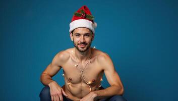 A portrait featuring a gay man seated against a blue backdrop adorned with Christmas lights and wearing a Santa Claus hat for Christmas Eve photo