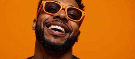 A bearded black man with orange glasses grins at the camera against an energetic orange backdrop photo