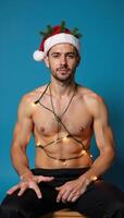 A portrait featuring a gay man seated against a blue backdrop adorned with Christmas lights and wearing a Santa Claus hat for Christmas Eve photo