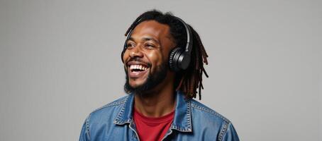 A happy African American man sporting a beard dreadlocks denim jacket and headphones is pictured against a grey backdrop listening to music in a studio setting photo