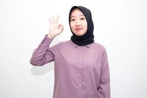 portrait of Indonesian Asian woman wearing shirt and hijab showing ok sign with one hand on eyes looking at camera through fingers. giving sign of agreement. Isolated on white Background photo