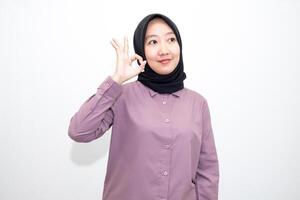portrait of Indonesian Asian woman wearing shirt and hijab showing ok sign with one hand on eyes looking at camera through fingers. giving sign of agreement. Isolated on white Background photo