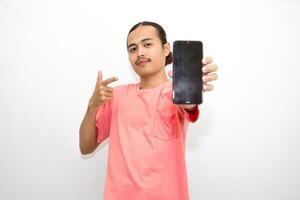 Asian, Indonesian man standing wearing pink shirt and holding mobile phone pointing to mobile phone and presenting using hand gesture. Isolated on white background photo