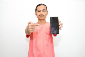 Asian, Indonesian man standing wearing pink shirt and holding mobile phone pointing to mobile phone and presenting using hand gesture. Isolated on white background photo
