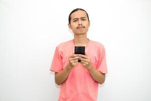 Asian, Indonesian man standing holding cellphone with hand on chin as if thinking hard, thinking about work or school assignment to find solution. Isolated on white background photo