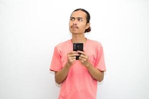 Asian, Indonesian man standing holding cellphone with hand on chin as if thinking hard, thinking about work or school assignment to find solution. Isolated on white background photo