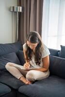 Young woman siting on the sofa and writing her personal diary, writing down thoughts, dreams and plans as part of psychotherapy after working with neurocoaching. High quality photo
