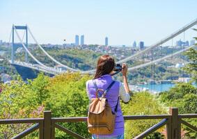 A young woman tourist with a backpack photographs the cityscape with a panoramic view of the Fatih Sultan Mehmet Bridge. Back view. Otagtepe Park photo