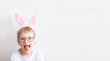 A boy in a bunny outfit. A happy child in glasses in a white t-shirt in rabbit ears shows the tongue on a white background. Happy Easter concept with place for text. photo
