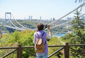 A young woman tourist with a backpack photographs the cityscape with a panoramic view of the Fatih Sultan Mehmet Bridge. Back view. Otagtepe Park photo