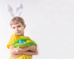 Happy child smiling in a yellow t-shirt in rabbit ears and with colorful eggs in a basket on a white background. Happy Easter concept with place for text. photo
