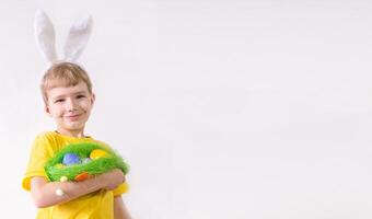 Happy child smiling in a yellow t-shirt in rabbit ears and with colorful eggs in a basket on a white background. Happy Easter concept with place for text. photo