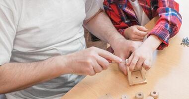 Happy father's day and childhood concept. Close-up of a father and a boy son in glasses work with hand tools, using a screwdriver, assembling a wooden house constructor at the table. photo