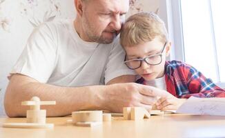 Happy father's day and childhood concept. Father and son-boy in glasses work with hand tools, using a screwdriver, assembling a wooden constructor at home at the table. photo