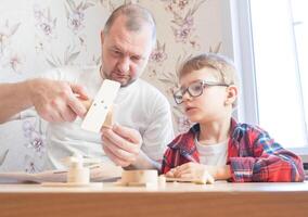 Happy father's day and childhood concept. Father and son-boy in glasses work with hand tools, using a screwdriver, assembling a wooden constructor at home at the table. photo