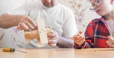 Happy father's day and childhood concept. Father and son-boy in glasses work with hand tools, using a screwdriver, assembling a wooden constructor at home at the table. photo