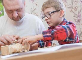 Happy father's day and childhood concept. Father and son-boy in glasses work with hand tools, using a screwdriver, assembling a wooden constructor at home at the table. photo