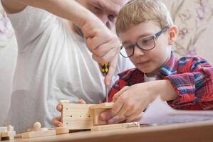 Happy father's day and childhood concept. Father and son-boy in glasses work with hand tools, using a screwdriver, assembling a wooden constructor at home at the table. photo