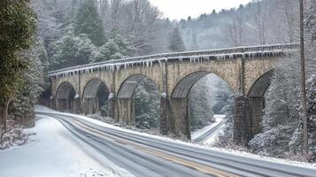Snowy winter landscape featuring a stone bridge over a winding road in a forest setting photo
