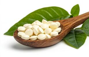 White capsules placed on a wooden spoon alongside green leaves against a plain background photo