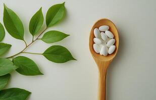 White capsules placed on a wooden spoon alongside green leaves against a plain background photo