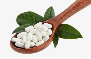 White capsules placed on a wooden spoon alongside green leaves against a plain background photo