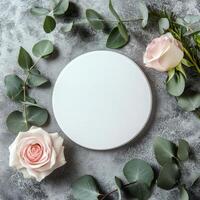 Circular blank canvas surrounded by pink roses and greenery on a textured gray surface photo