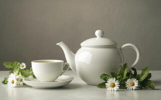 A white teapot with a cup and daisies, set on a light backdrop with green mint leaves photo