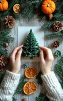 Making a festive autumn paper tree. A person assembles a green paper Christmas tree surrounded by autumn foliage, dried fruit, and pinecones on a wooden surface. photo