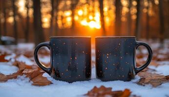 Two cups of coffee are sitting on a snowy ground with leaves in the background. The cups are black and have a star pattern on them. The scene is peaceful and serene photo