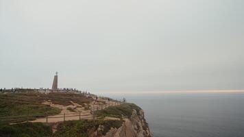 Portugal, Lisbon - July 29, 2022. Cape with tourists and cross. Action. Memorial cross at cliff with sea. A lot of tourists at Cape Rock with monument on cloudy day video