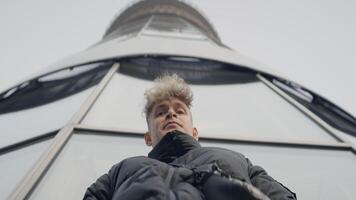 Bottom view of young man on background of tower. Action. Stylish young man in jacket at modern tower. Young man looks around warily video