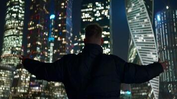 Man raises hands to glowing buildings. Action. Man with hands raised to cityscape. Man stands on background of glowing modern skyscrapers at night video