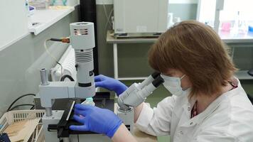 Woman looks through microscope in laboratory. Stock footage. Masked woman looks at biochemical substance through microscope. Analysis of substance under microscope in laboratory video