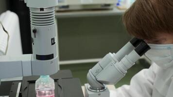 Woman looks through microscope in laboratory. Stock footage. Masked woman looks at biochemical substance through microscope. Analysis of substance under microscope in laboratory video