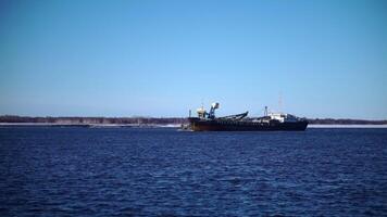 View of floating large vessel with cranes . Clip. Cargo ship with cranes floats on river on sunny day. Cargo ship sails on sunny winter day on background of snowy shore video