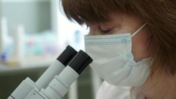 Woman looks through microscope in laboratory. Stock footage. Masked woman looks at biochemical substance through microscope. Analysis of substance under microscope in laboratory video