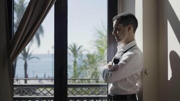 Side view of a man in white suit standing by the window. Action. Sea and palm trees behind the window. video
