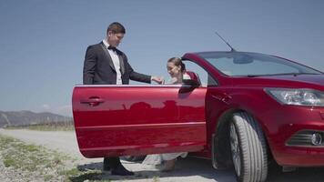 Newlyweds by the convertible car, feelings of tenderness. Action. Man in suit helps the bride to leave the red car. video