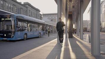Serbia, Belgrade - April 12, 2023. Young man is talking on phone at bus stop. Action. Young man is waiting for bus at bus stop on sunny day. Modern electric buses on city routes video