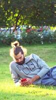 A young man is browsing on his smartphone while lying on nature on green grass in the summertime. Concept. Serious man using mobile phone while relaxing in a city park, lying on the grass, chatting video