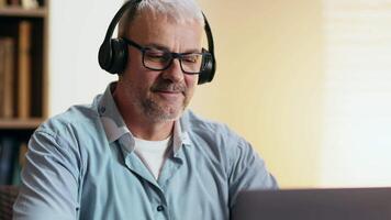 A senior man focused on his laptop while listening with headphones in a sunny, inviting home environment. video
