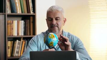 A man explores a globe while working on a laptop in a warm, inviting space filled with books, teacher gives online lesson video